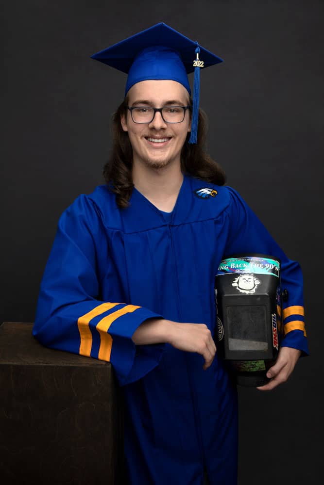 Senior photo of a smiling young man with long brown hair. He's wearing a bright blue cap and gown and holding a welding helmet. Max Anderson Senior Portrait Photography.