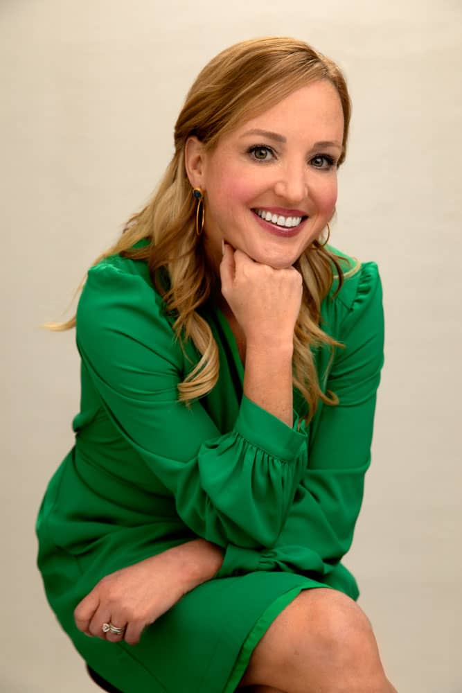 Professional photo of a pretty, smiling young woman with dirty blonde hair and red lipstick. She's wearing a green dress while sitting on a stool with legs crossed, arms resting on her legs and chin resting on her fist. Meredith Dowless Personal Branding Photography.