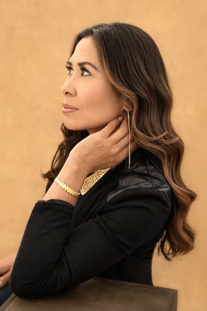 Bright photo of a pretty woman sitting and leaning on a box, her hand up as she touches her neck. She is wearing a black shirt with golden earrings, necklace and a bracelet. Myra Ruiz Personal Branding Photography.