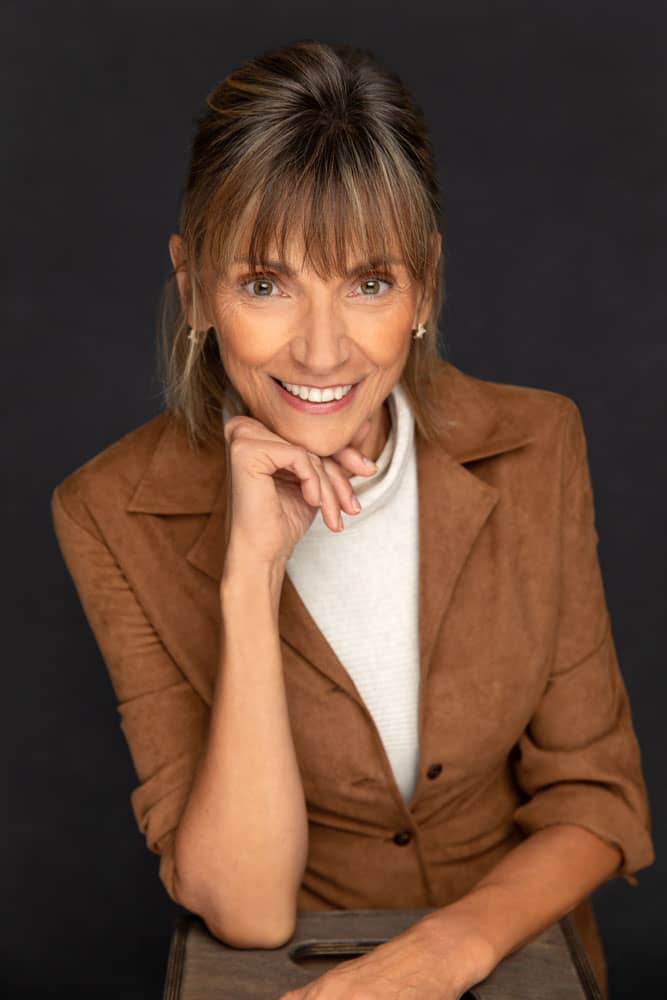 Professional photo of a pretty, smiling woman with dirty blonde hair and hazel eyes leaning on a box with her chin resting on her hand. She's wearing a white turtle neck and a brown jacket. Regina Yorke Headshot Photography.