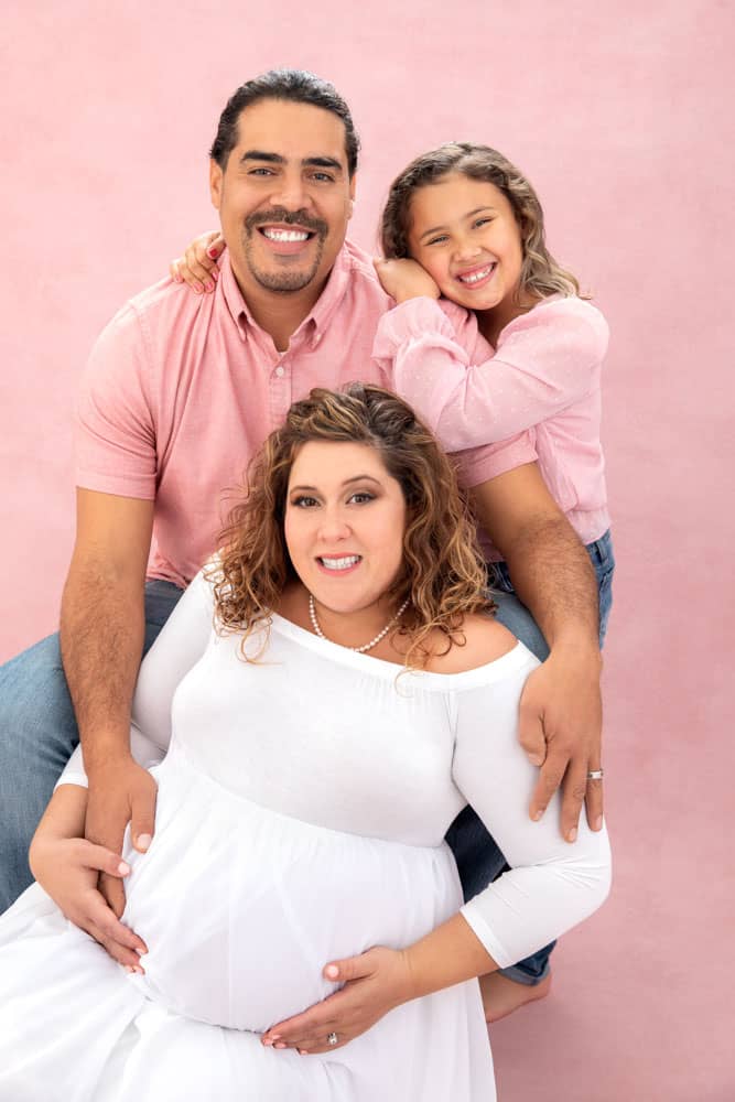 Precious family and maternity photo of father, mother, daughter and baby on the way. Mother is wearing a white dress and pearls, father and daughter are wearing pink shirts. Jessica Stellato-Pagan Maternity and Family Portrait Photography.