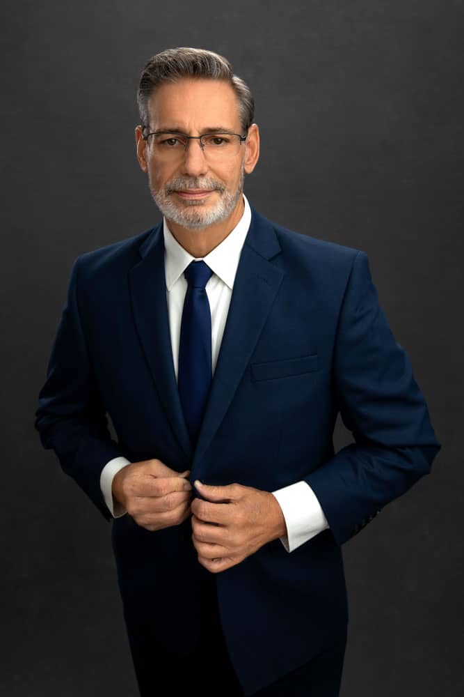 Sharp photo of a handsome man in a blue suit, white shirt and blue tie with a salt and pepper beard. He's buttoning his suit jacket and looking serious. Donald Aucoin personal branding photography.