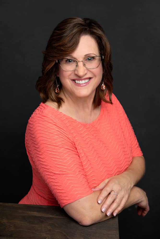 Nice headshot of a pretty, smiling woman with coral shirt on a dark backdrop. She's leaning on a box and folding her arms. Charlotte Aucoin Personal Branding Photography
