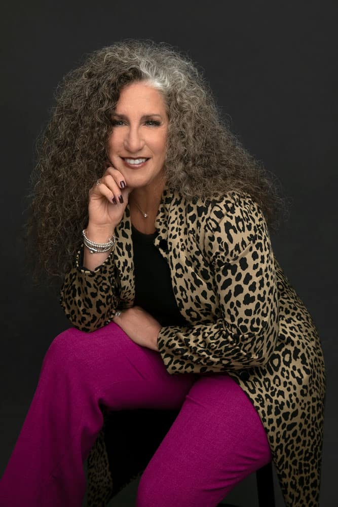 Classy photo of a pretty, smiling woman with pretty wavy salt and pepper hair. She's wearing a cheetah print jacket and hot pink pants on dark backdrop. Cinnamon McCauley Women's Portrait Photography.