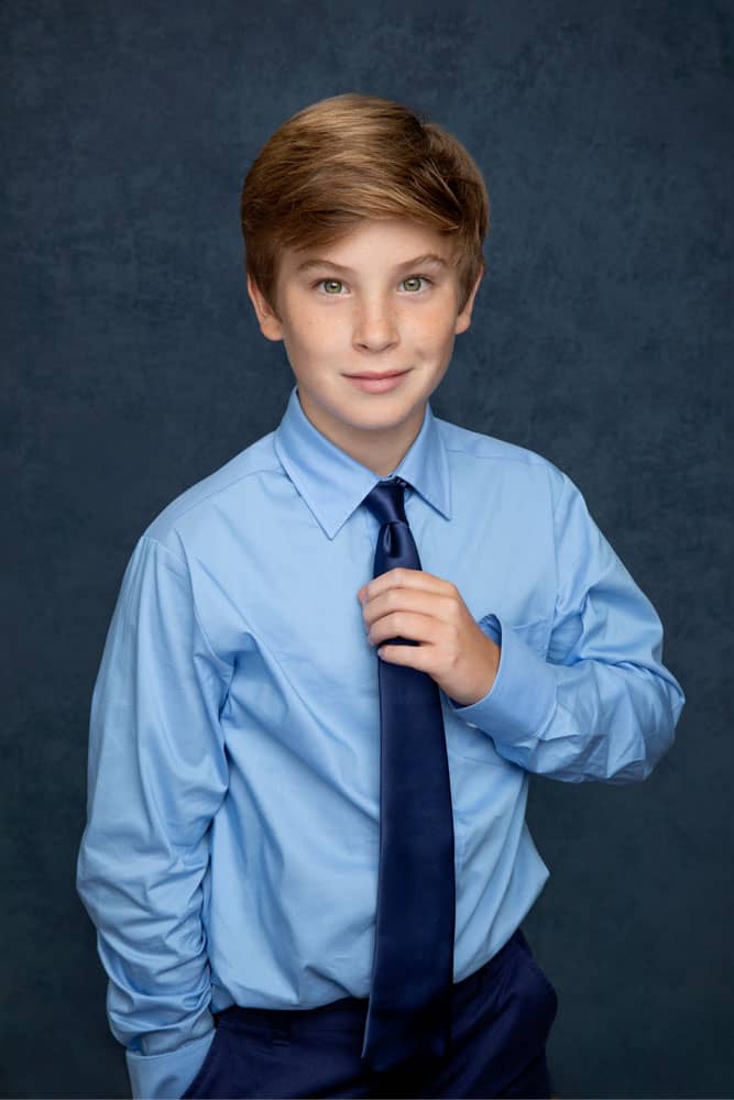 Cute photo of a handsome young man with a smirk, wearing a light blue shirt with a dark blue tie. He's holding his tie with one hand. Guy Furay and Myra Ruiz Family Portrait Photography.