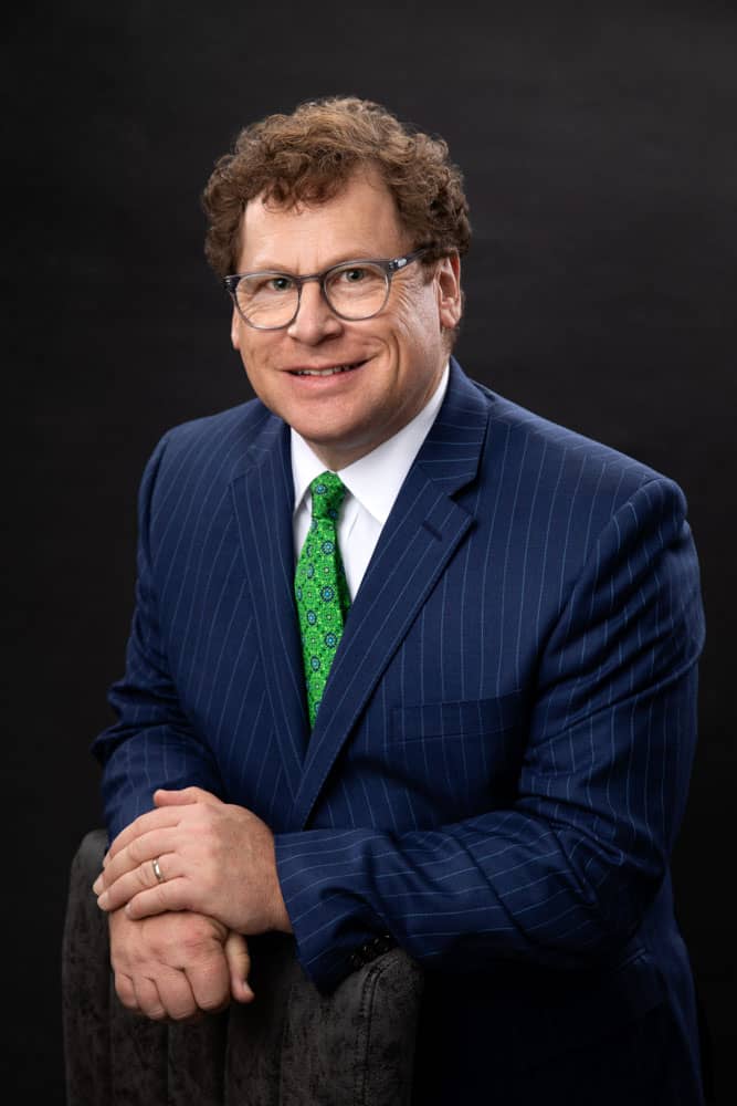 Sharp photo of smiling Guy Furay with curly brown hair in a blue pinstripe suite, white shirt and bright green tie. He's casually leaning on a chair. Guy Furay Personal Branding Photography.