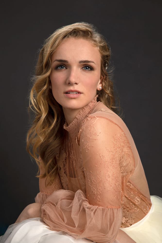 Elegant photo of a pretty young woman with long dirty blonde hair in a peach colored shirt with a white toole skirt. She has a slight smirk and is wearing pearl earrings. Holly Yurkin Women's Portrait Photography.
