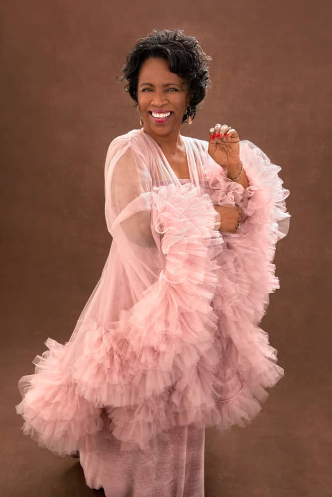 Fun photo of a pretty, smiling black woman with glasses wearing a pink tulle dress and golden earrings. She's wearing red lipstick and red nails. Janice Butler Women's Portrait Photography.