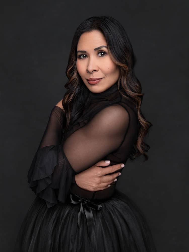 Moody photo of strong, pretty woman with long brown hair and red lipstick. She's standing and leaning back, her arms wrapped around her torso. She's wearing a black mesh shirt with a black toole skirt. Myra Ruiz Personal Branding Photography.
