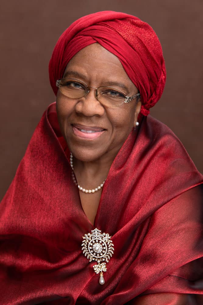 Classy photo of a pretty black woman smiling, wearing pearl earrings and a necklace. She's wearing a red head covering and a red sash connected with a pretty brooch, also a pearl necklace and earings. Photo has rich colors with the red on a brown backdrop. Audra Odom Women's Portrait Photography.