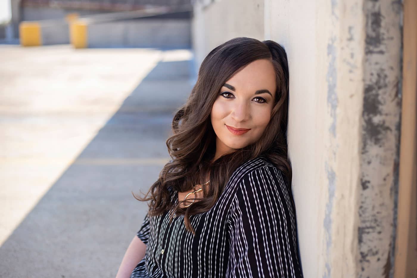 Senior photo of a pretty young woman with long brown hair and red lipstick. She's wearing a striped shirt and leaning on textured wall in a parking garage. Chelsea Bergeron Senior Portrait Photography.