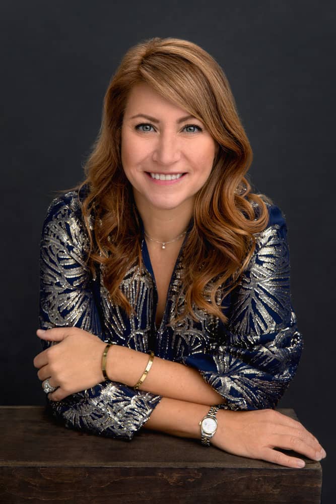 Classy photo of a pretty, smiling woman with brown hair leaning on a box, holding her arms. She's wearing a nice blue shirt with gold florals. She's wearing gold bracelets and a watch on her wrist. Chris and Amber Edwards Family Portrait Photography.
