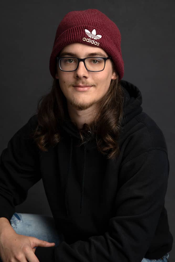Senior photo of a smirking young man with long brown hair. He's wearing a red Adidas beanie and black hoodie. Max Anderson Senior Portrait Photography.