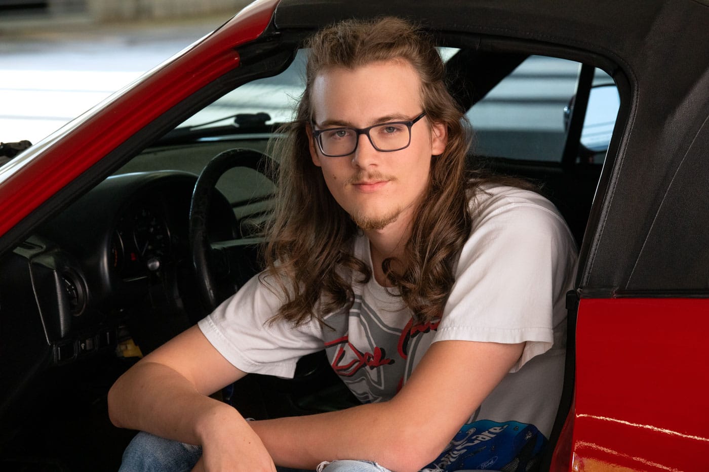 Senior photo of a young man with long brown hair and glasses. He's wearing white graphic tee as he sits in his red 1990 Mazda Miata. Max Anderson Senior Portrait Photography.