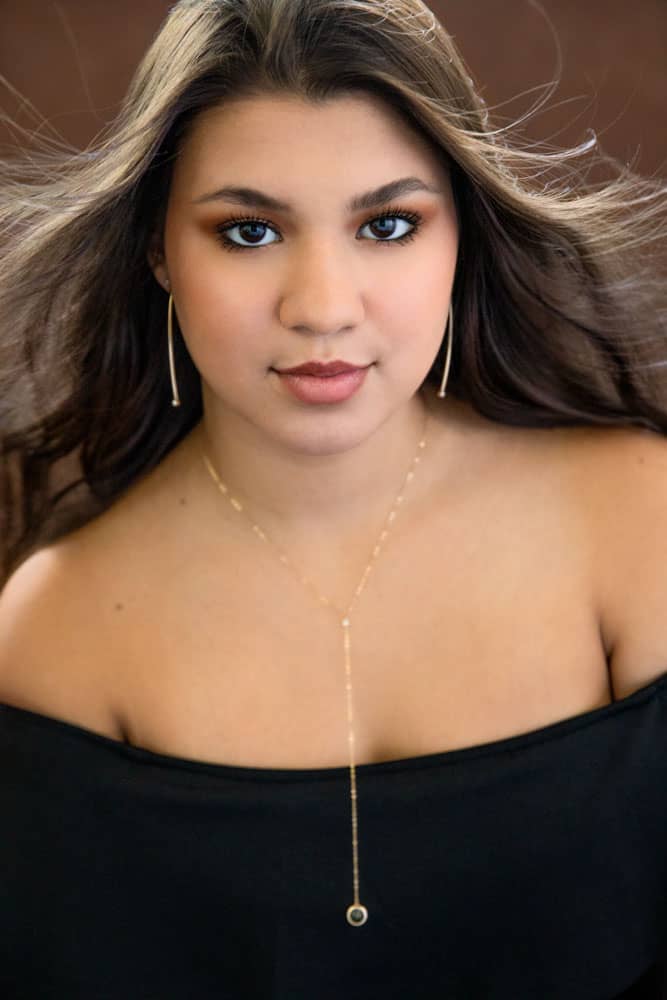 Classy senior photo of a pretty young woman with long brown hair and dark brown eyes. She's wearing gold earrings, a gold necklace and a black dress. Her hair is blowing in the wind. Nicole Calderon Senior Portrait Photography.
