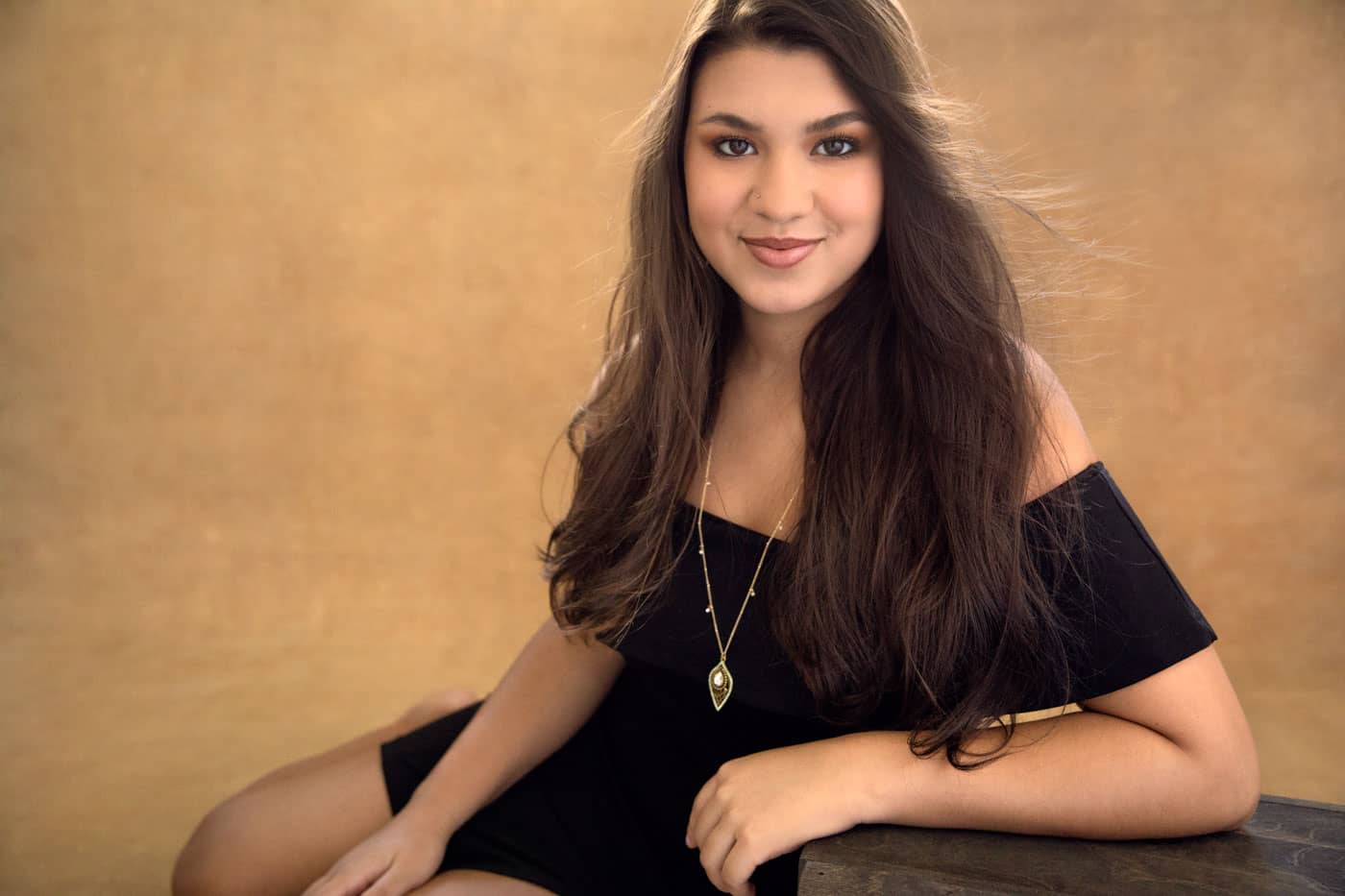 Classy senior photo of a pretty young woman with long brown hair and dark brown eyes, sitting and leaning on a box. She's wearing a necklace with a gold pendant and a black dress. Nicole Calderon Senior Portrait Photography.