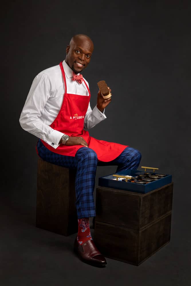 Branding photo of a stylish, handsome black man in bright red apron with red socks and bowtie. He is sitting on a box, with his shining tools in front of him and is ready to shine leather shoes. Charles Davis of A Polished Man during personal branding photo session.