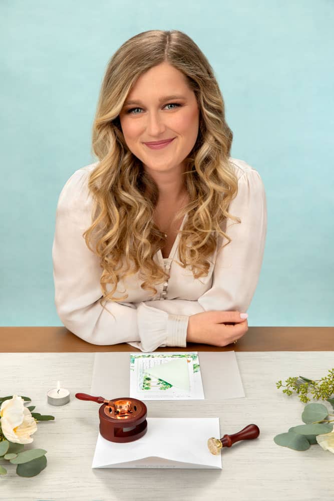 Branding photo of a pretty, smiling young woman with blond hair and creme shirt leaning on table. She is sealing a wedding invitation with wax. Personal branding photography session with Printed Water and Alicia Waller.