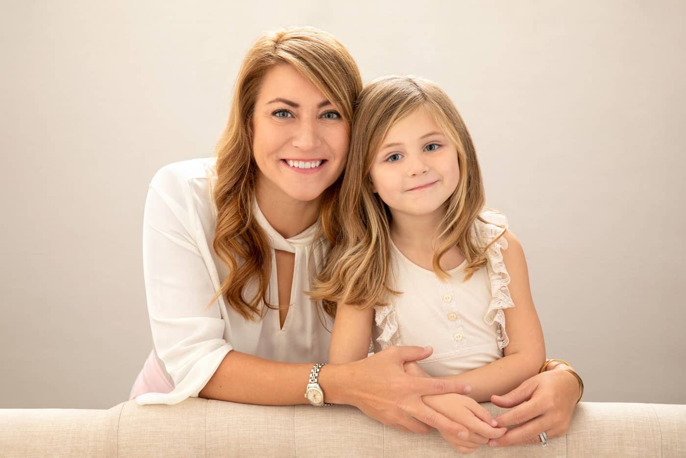 Precious photo of a beautiful mother and daughter standing close and leaning on a couch. The mother, who has long dirty blonde hair, has her arms around her daughter. They both are wearing white shirts. Chris and Amber Edwards Family Portrait Photography.