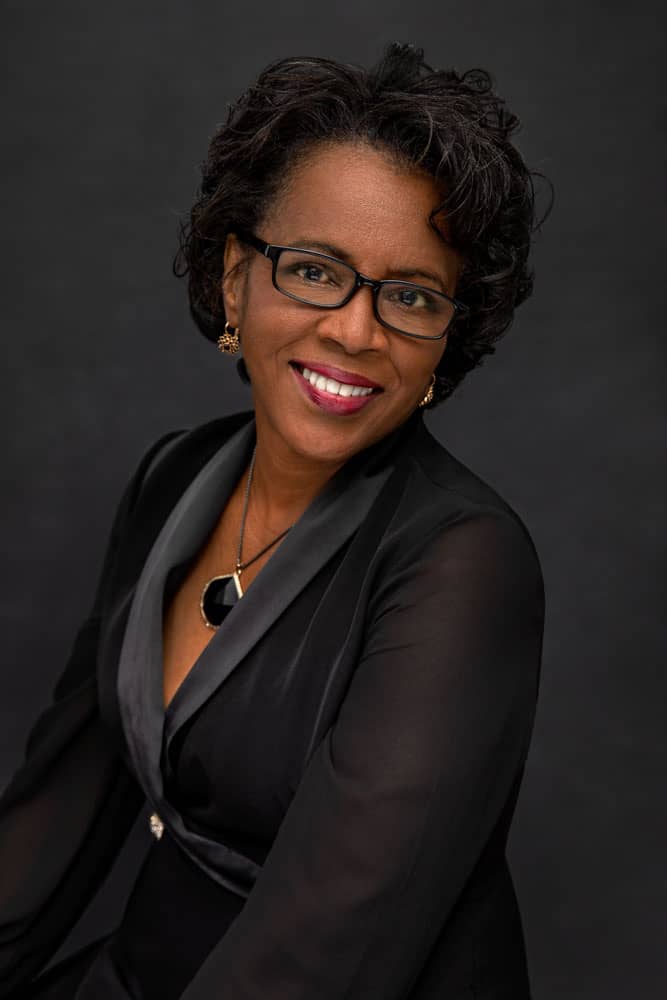 Classy photo of a pretty, smiling black woman with glasses wearing red lipstick, a black shirt, black necklace and golden earrings. Janice Butler Women's Portrait Photography.