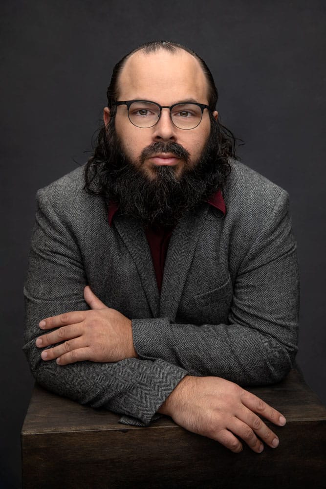 Sharp photo of an actor playing a sophisticated professor or lawyer. Wearing a red shirt with a gray jacket. His hair slicked back and his beard tamed. Jonathan Hano Acting and Headshot Photography.
