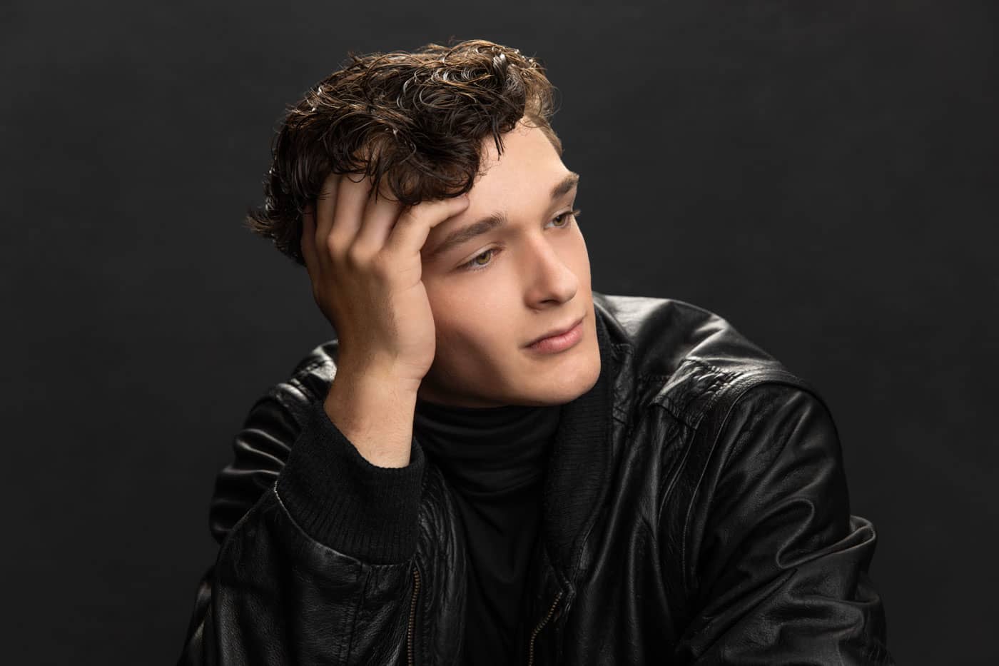 Sharp photo of a handsome young man with curly brown hair looking away wearing a black leather jacket. His head is leaning on his hand, with his fingers in his hair. Lucas Nichter Senior Portrait Photography.