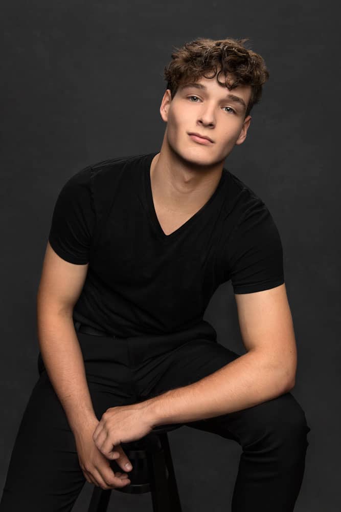 Sharp photo of a handsome young man with curly brown hair sitting on a stool and leaning on his knee wearing a black v-neck shirt. Reminds us of Patrick Swayze. Lucas Nichter Senior Portrait Photography.