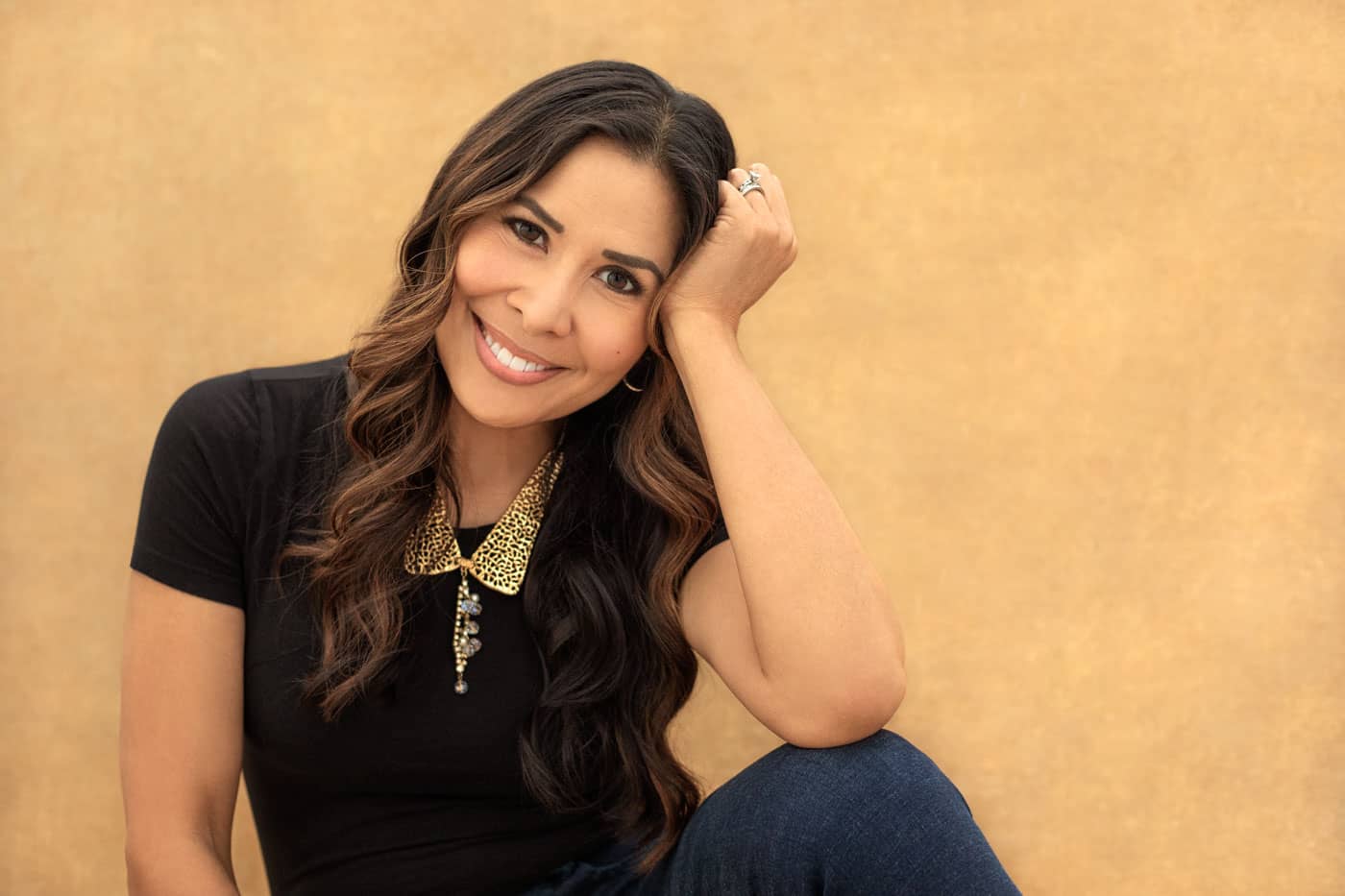 Bright branding photo of a pretty, smiling woman sitting with her elbow resting on her knee and her head rested on her hand. She's wearing a black shirt, jeans and a gold necklace. Myra Ruiz Personal Branding Photography.