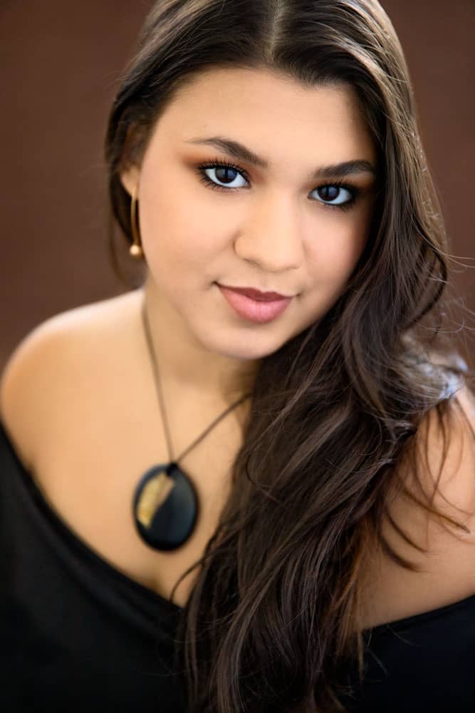 Classy senior photo of a pretty young woman with long brown hair and dark brown eyes. She's wearing a necklace with a black pendant and a black dress. Nicole Calderon Senior Portrait Photography.