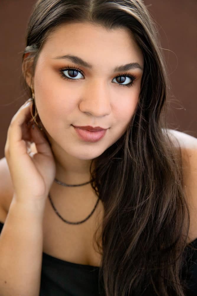 Classy senior photo of a pretty young woman with long brown hair and dark brown eyes. She's wearing a black necklace and a black dress. Her hand is touching her neck. Nicole Calderon Senior Portrait Photography.