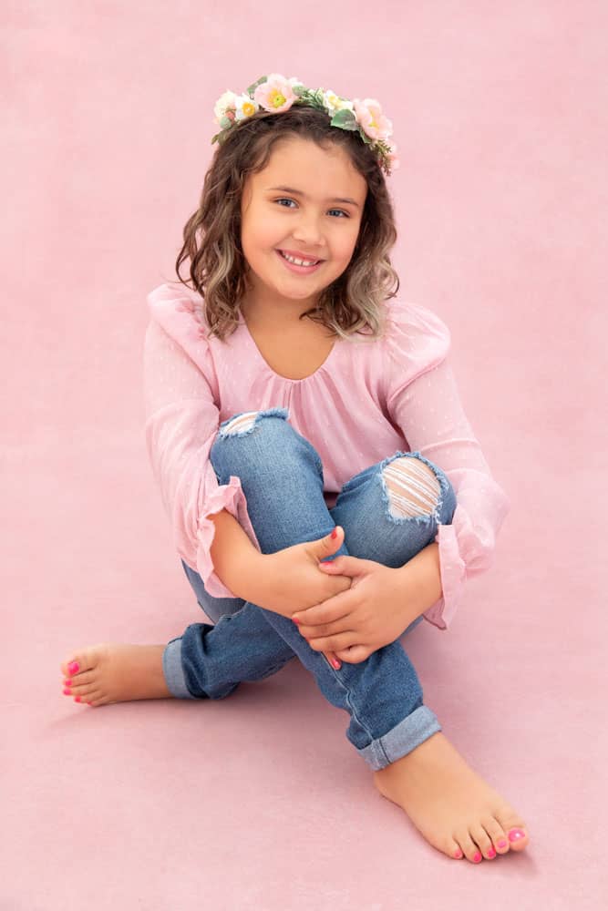 Precious photo of adorable little girl with brown hair. She's sitting on the floor, hugging her knees. She's wearing a pink shirt, torn jeans and a flower crown. Jessica Stellato-Pagan Maternity and Family Portrait Photography.