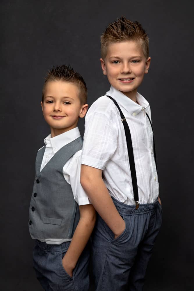 Cute photo of two young brothers standing back to back and smiling. Both are wearing white shirts and gray pants. One is wearing a gray vest and one is wearing black suspenders. Family Portrait Photography.