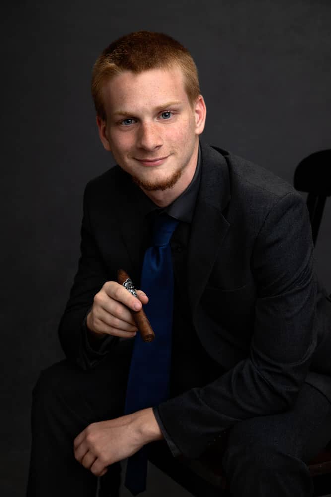 Senior photo of a young man graduating from Greenville High School. He is wearing a black shirt, black jacket and pants with a blue tie. He is sitting in a chair, leaning forward and holding a cigar, a family coming-of-age tradition. Tristan Kopplin Senior Portrait Photography.
