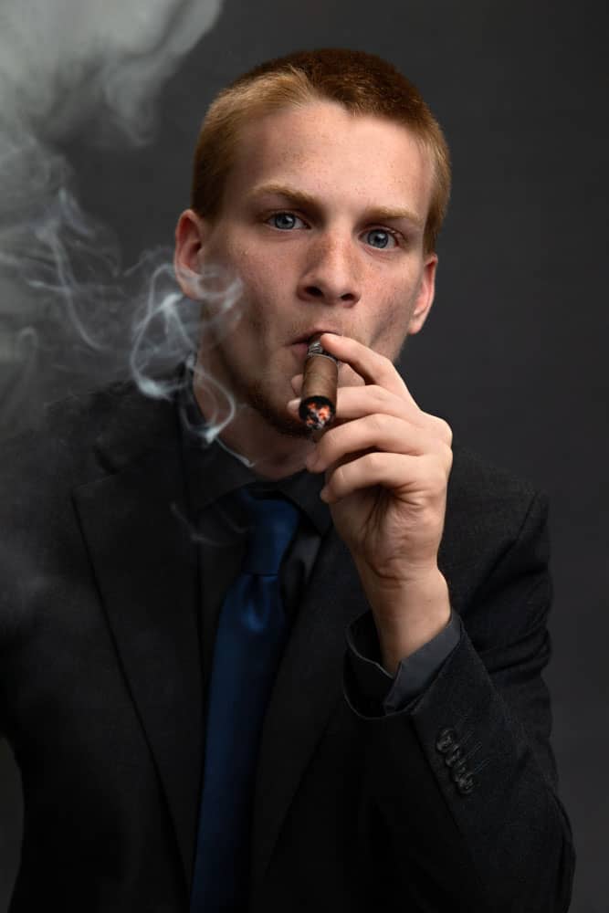 Senior photo of a young man graduating from Greenville High School. He is wearing a black shirt, black jacket and pants with a blue tie. He is standing and smoking a cigar, which is a family coming-of-age tradition. Tristan Kopplin Senior Portrait Photography.