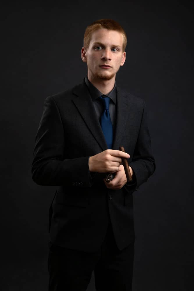 Senior photo of a young man graduating from Greenville High School. He is wearing a black shirt, black jacket and pants with a blue tie. He is standing, looking away and holding a cigar, a family coming-of-age tradition. Tristan Kopplin Senior Portrait Photography.