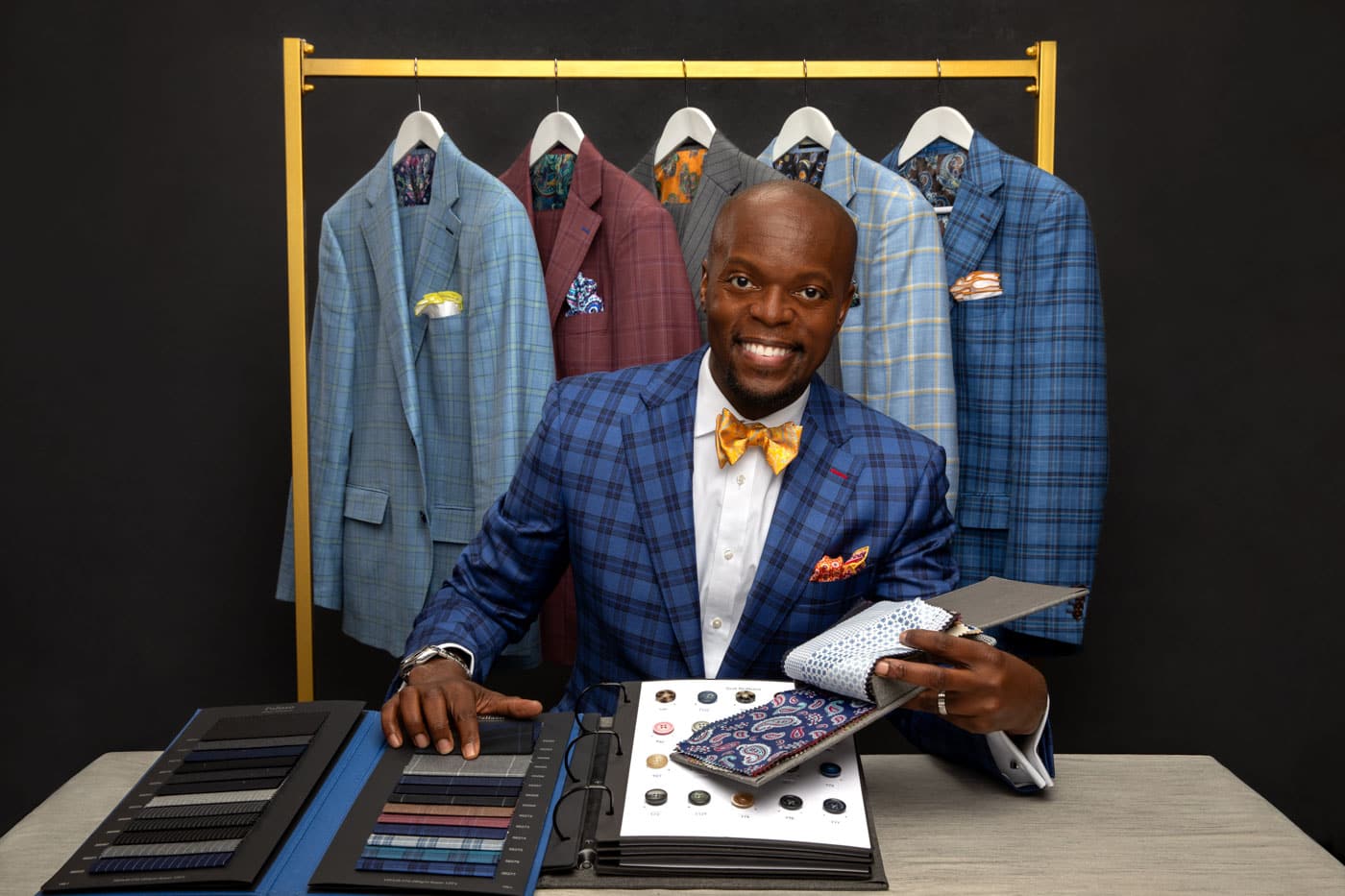 Product photo of a stylish, handsome black man in dark blue suit with paisley bowtie, floral pocket square, and yellow socks. He's sitting at a table, showing you the patterns of his products. A rack of suits hangs behind him. Charles Davis of A Polished Man during personal branding photo session.