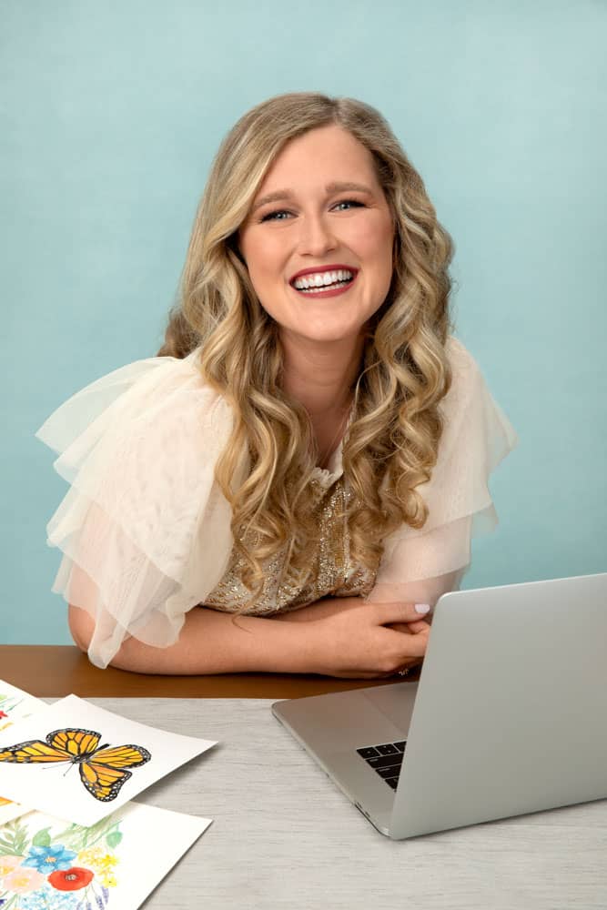 Branding photo of a smiling pretty young woman with blond hair and cream tulle leaning on table working on laptop. Personal branding photography session with Printed Water and Alicia Waller.
