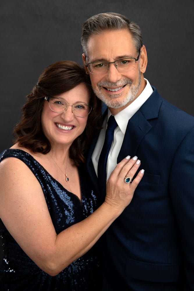 Cute engagment photo of couple standing and holding each other. Attractive woman with glasses in a blue sequin dress has her hand on her fiance's chest. Attactive man with glasses in a blue suit with blue tie and white shirt. Engagement portrait photography.