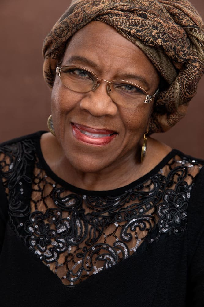 Classy photo of a pretty black woman smiling. She's wearing a brown, textured head covering and a black shirt on a brown backdrop. Audra Odom Women's Portrait Photography.