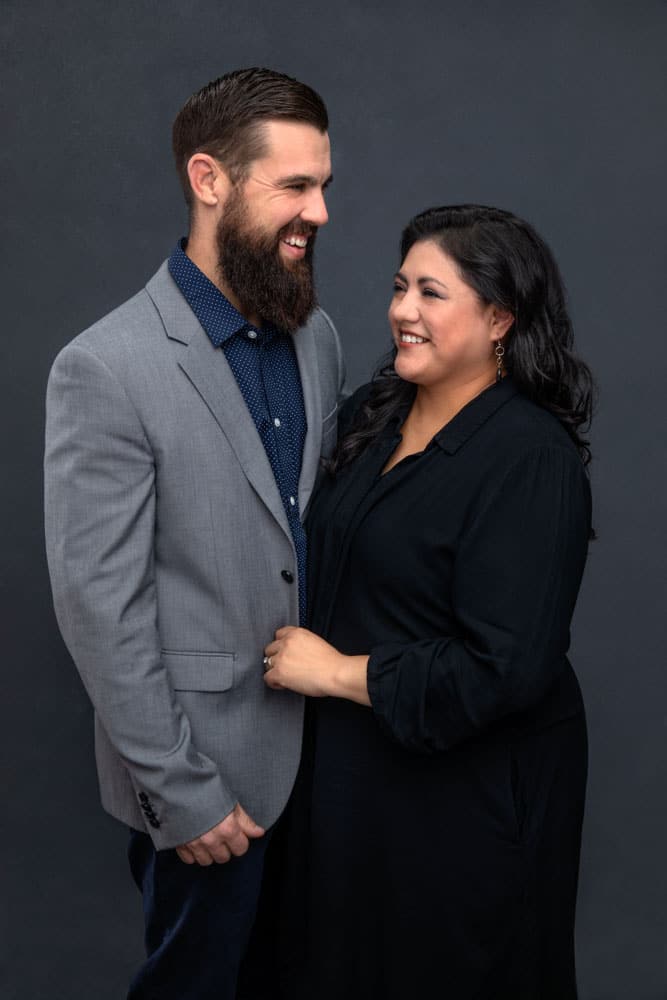 Precious photo of a cute, smiling couple standing close and looking at each other. He is wearing a blue shirt and gray jacket. She is wearing a black dress. Her hand has a ring and is pulling on his jacket. Josh and Stefanie Fraley Family Portrait Photography.