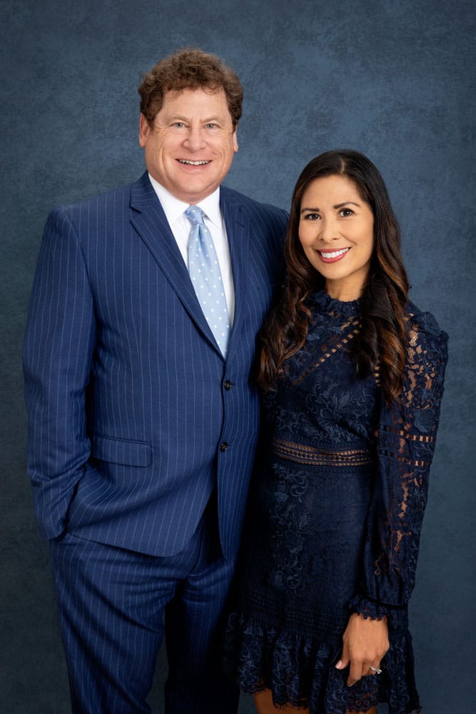Precious photo of a cute husband and wife standing close, their arms around each other's backs. Guy is wearing a striped blue suit and Myra is wearing a dark blue dress. Guy Furay and Myra Ruiz Family Portrait Photography.