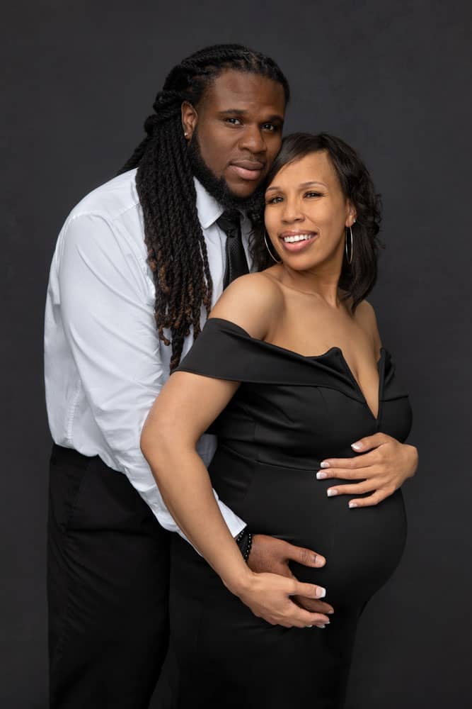 Precious maternity photo of cute husband and wife standing close and holding each other. He is standing behind her with his hand under her baby belly. She's holding her baby belly with one hand and holding his hand with the other hand. Holmes Family Portrait Photography.
