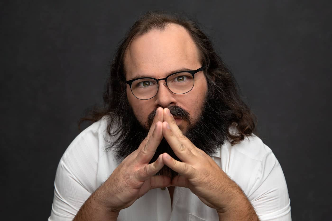 Branding photo of an actor playing a clever character with evil intent. Wearing a white shirt and black glasses, his fingers together covering his mouth. Jonathan Hano Acting and Headshot Photography.