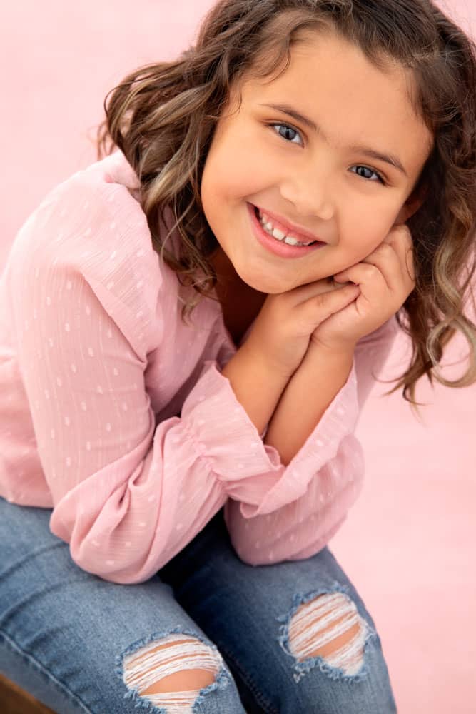 Cute photo of an adorable little smiling girl with brown hair. She's sitting with her elbows on her knees and her face resting on her hands. She's wearing a pink shirt with torn blue jeans. Jessica Stellato-Pagan Maternity and Family Portrait Photography.