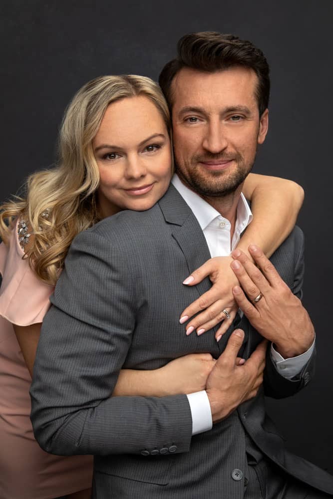 Cute photo of a married couple close together, the blonde haired wife is standing behind the brown haired husband with her arms around him. He is wearing a gray suit and white shirt. She is wearing a pink dress. Family Portrait Photography.
