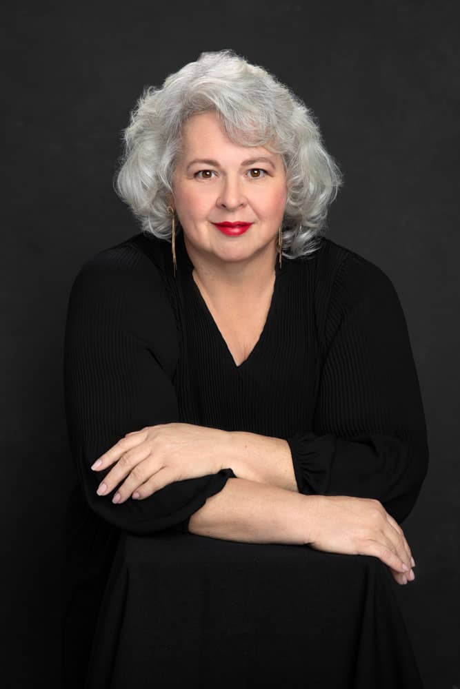 Elegant photo of a beautiful, smirking woman. She has beautiful silver hair and red lipstick. She is wearing a black shirt and leaning forward, arms folded over each other. Veronica Pitre Women's Portrait Photography.